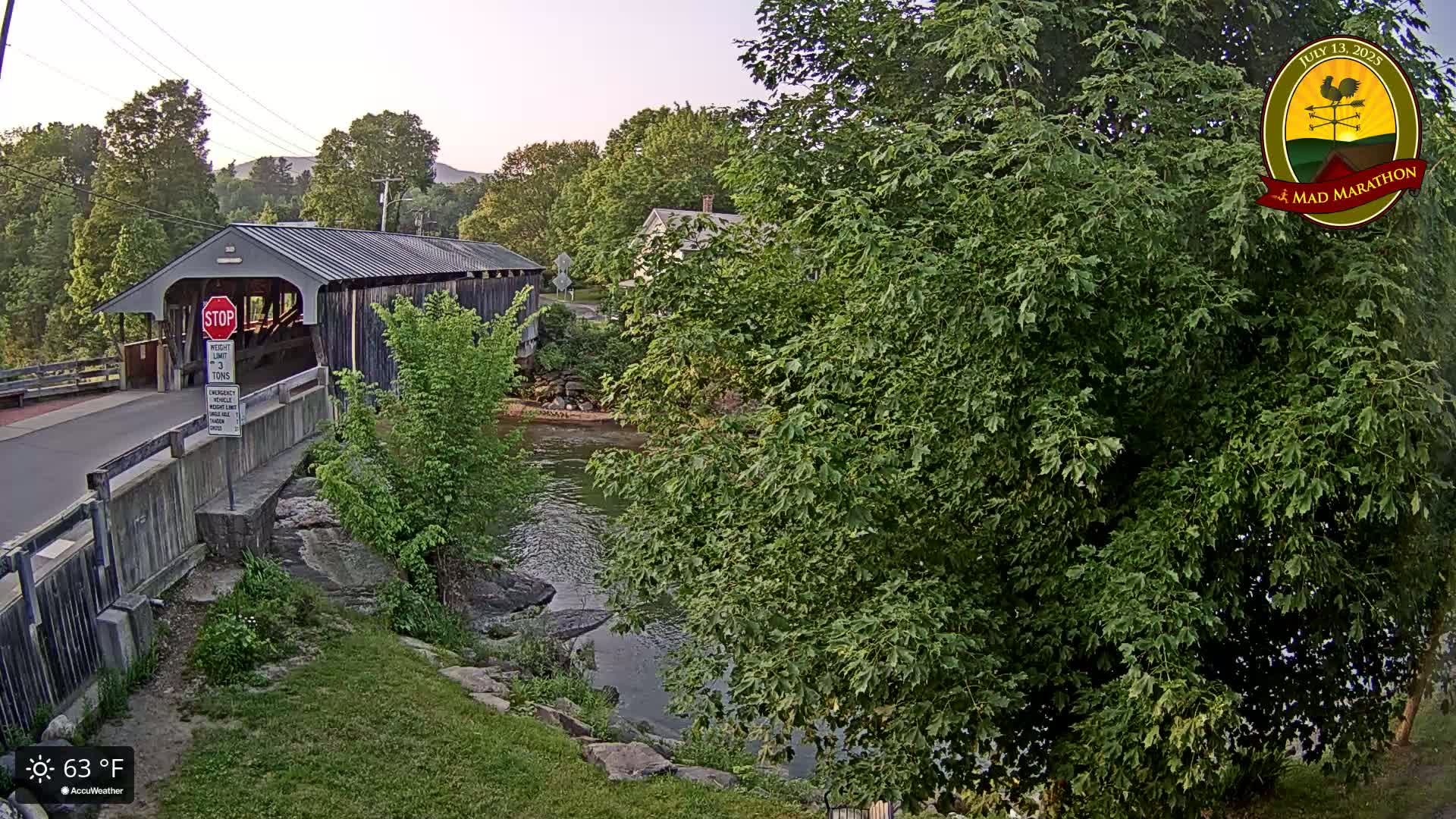 A covered bridge spans a river under a partly sunny sky, with lush green foliage surrounding the scene and the temperature at 63°F.