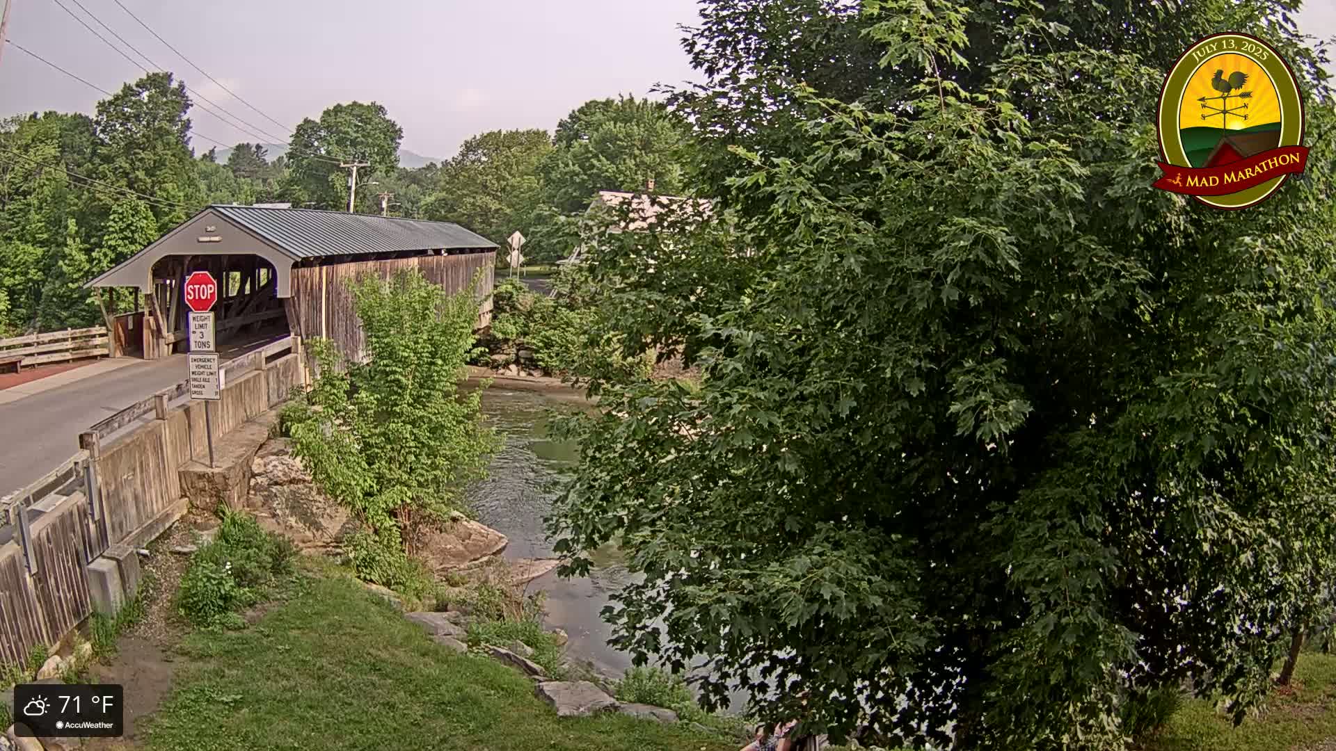 A covered bridge spans a calm river, surrounded by lush green trees and grassy banks under a partly cloudy sky.