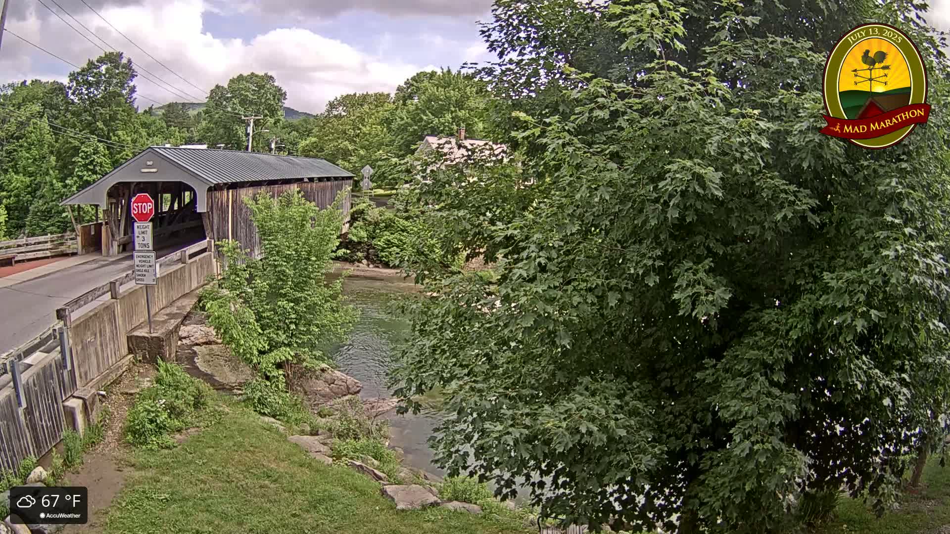 A covered bridge spans a calm river under a partly cloudy sky, surrounded by lush green trees and foliage.