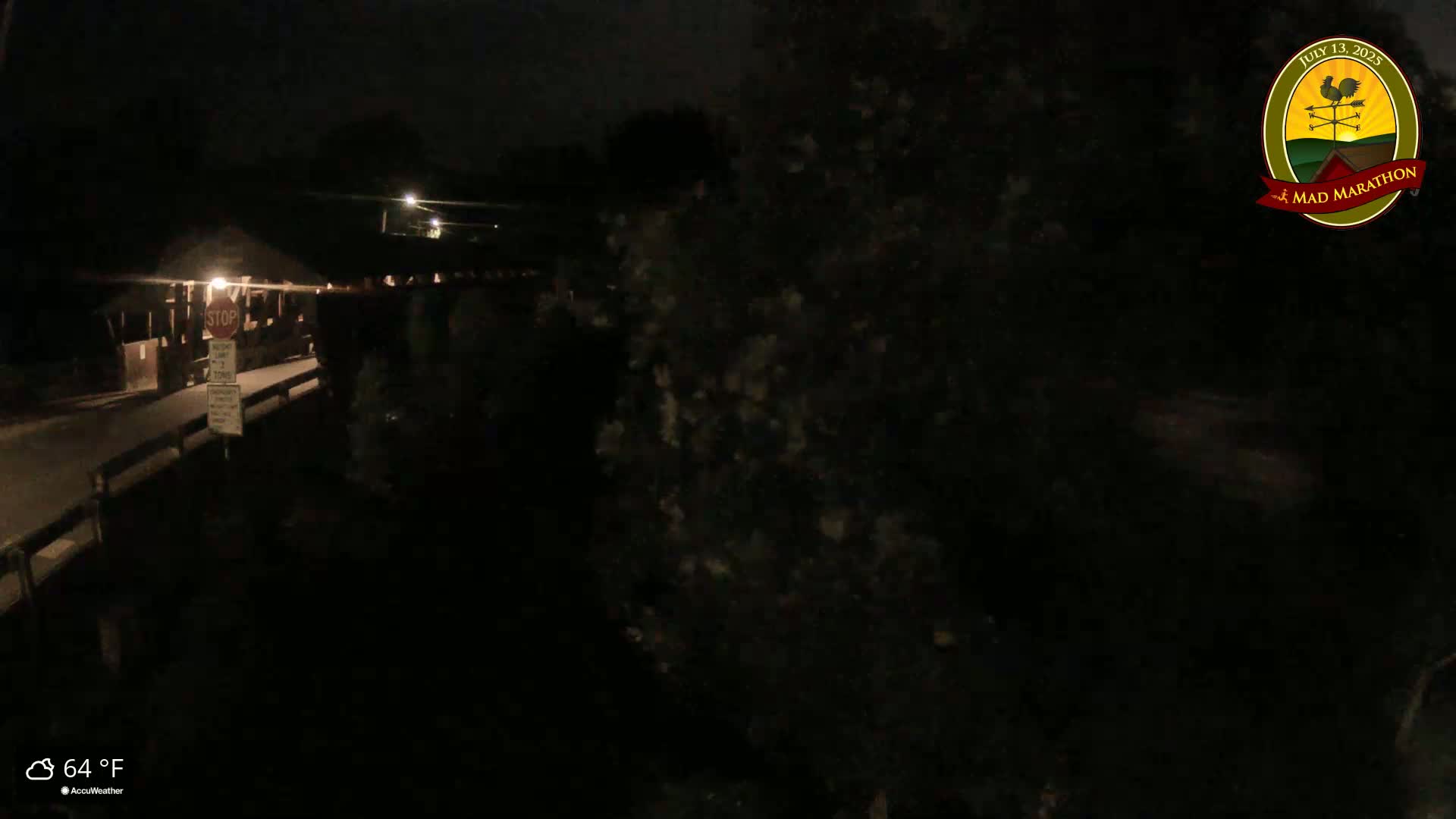 A nighttime view of a dark, empty road leading to a covered bridge under a clear, 64°F sky.