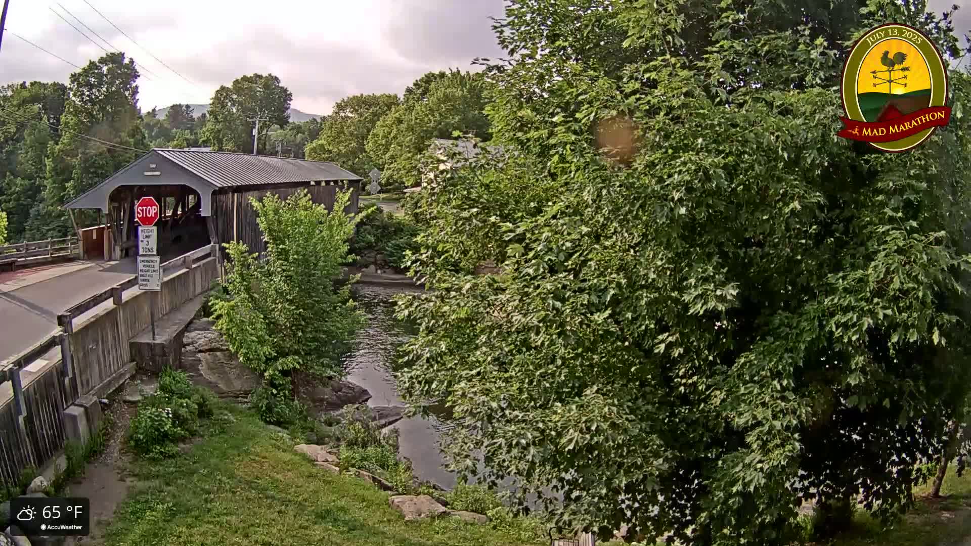 A covered bridge spans a calm river under a partly cloudy sky, surrounded by lush green trees and vegetation.