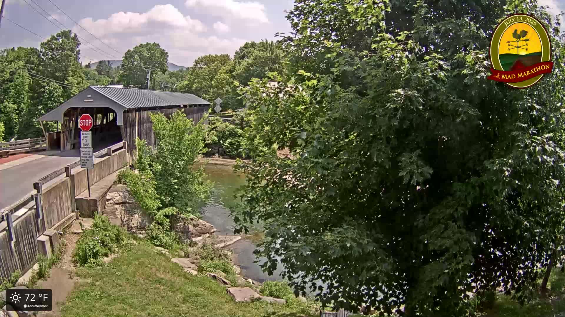 A covered bridge spans a calm river under a partly cloudy sky, surrounded by lush green trees and grassy banks.