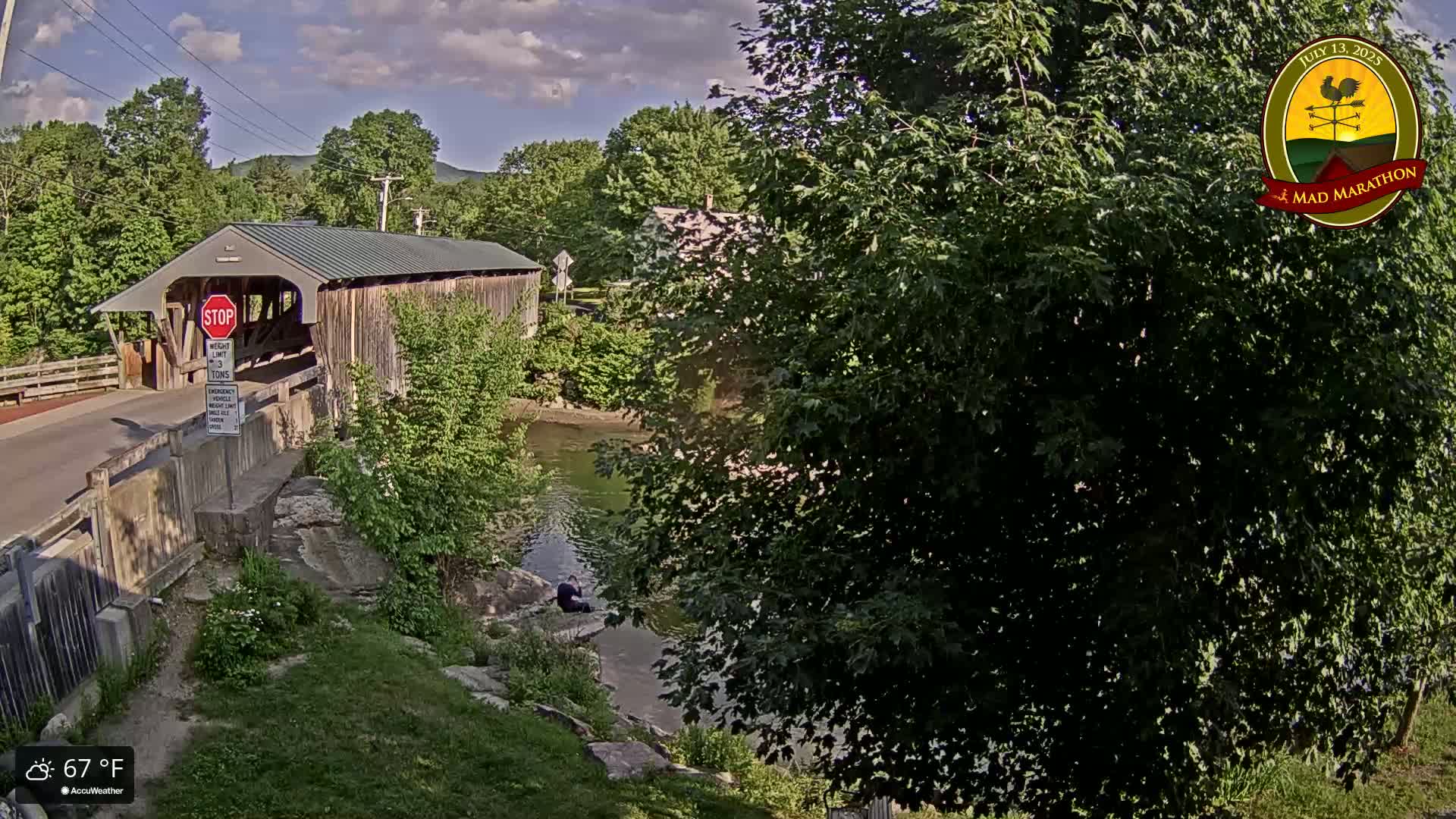 A covered bridge spans a calm river under a partly cloudy sky, surrounded by lush green trees and grassy banks.