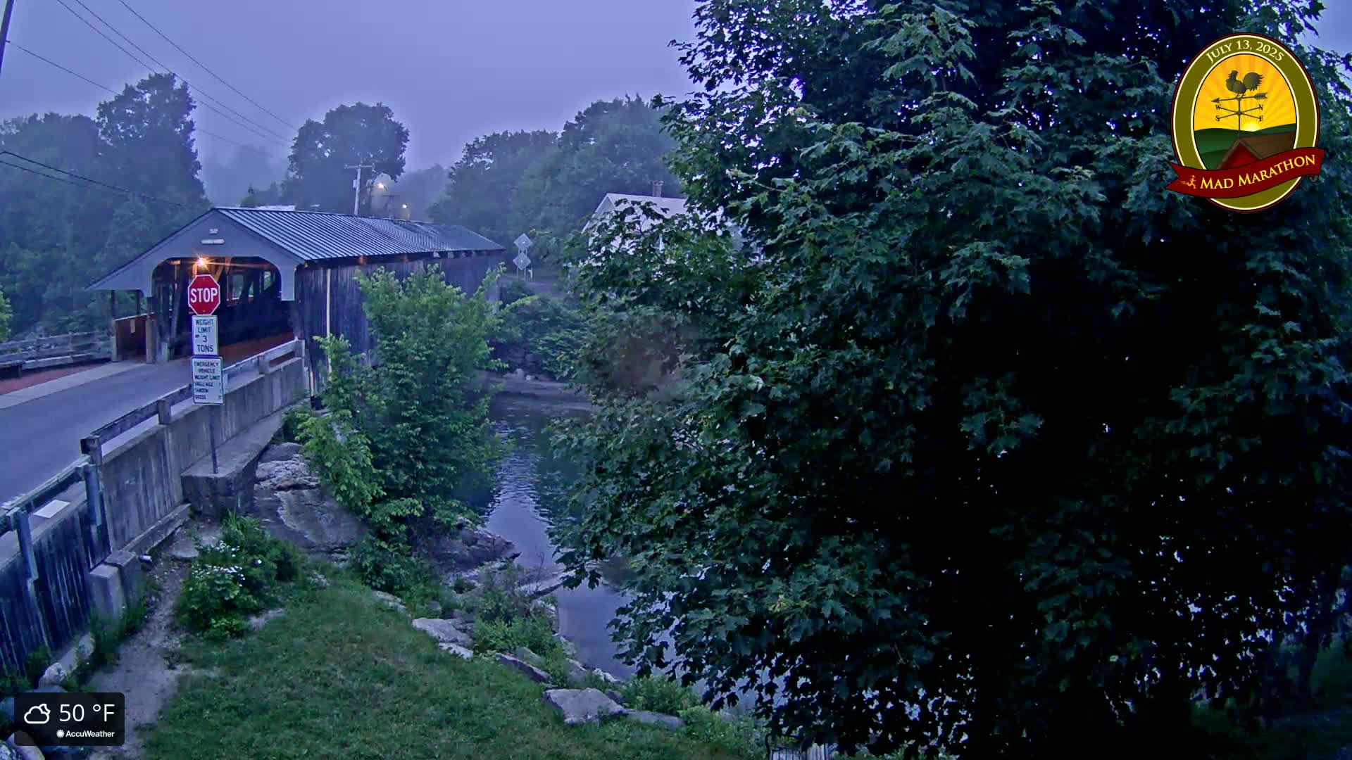 A covered bridge spans a calm river on a foggy, 50°F morning, surrounded by lush green foliage.