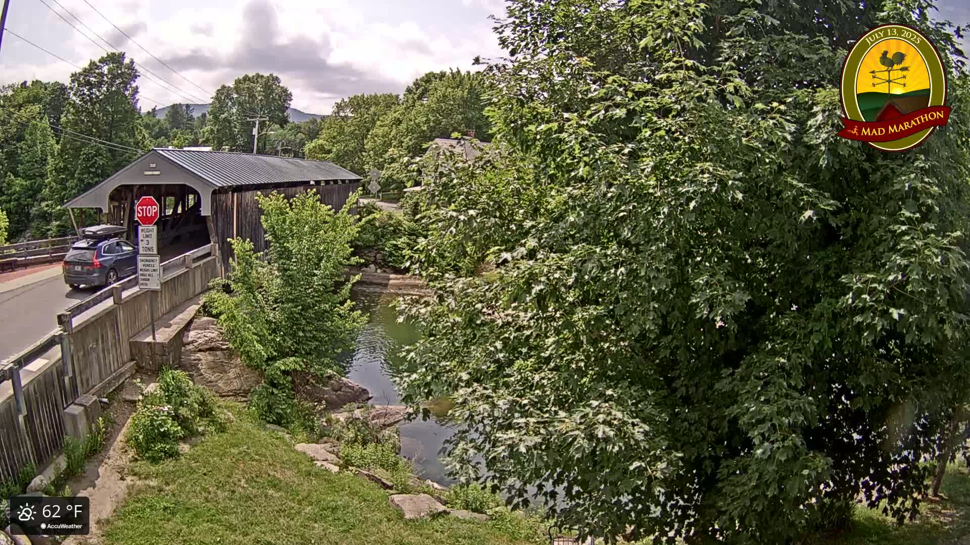 A covered bridge spans a calm body of water under a partly cloudy sky, with lush green foliage surrounding the scene.
