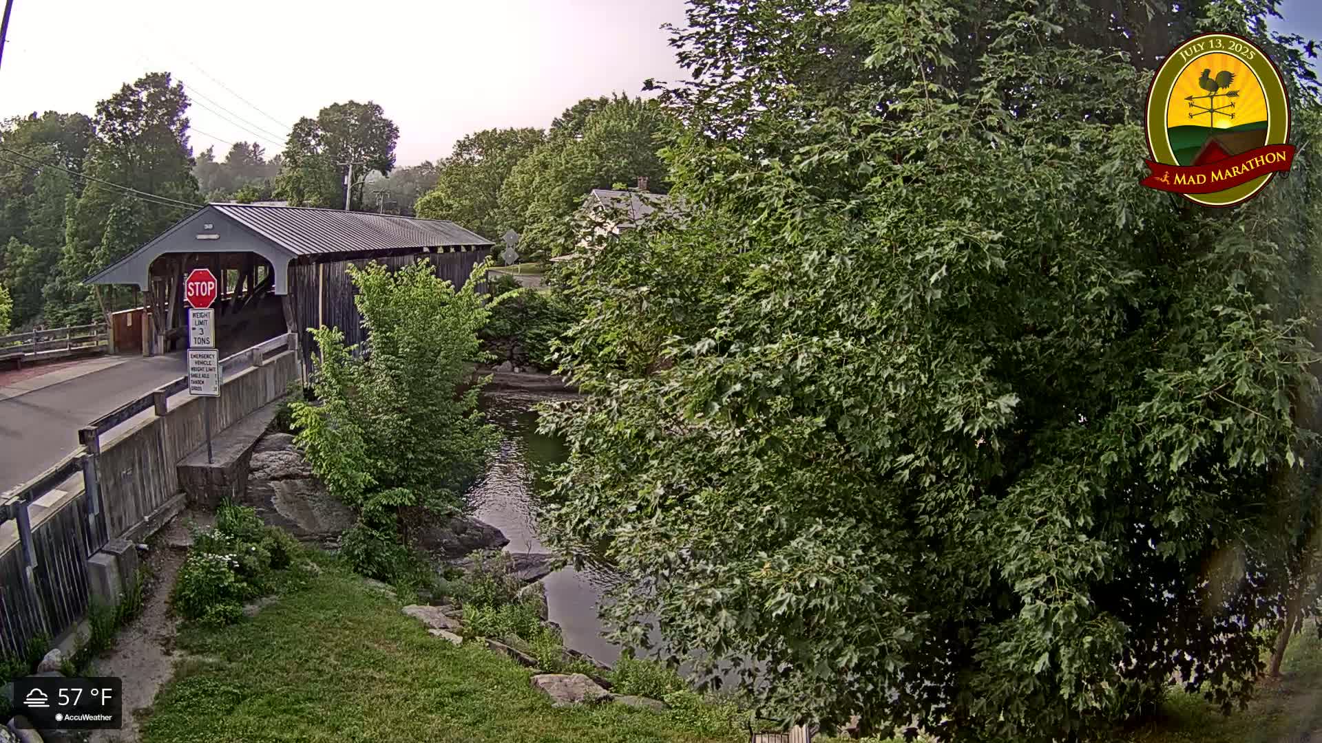 A covered bridge spans a calm river under a mostly cloudy sky, surrounded by lush green trees and foliage.