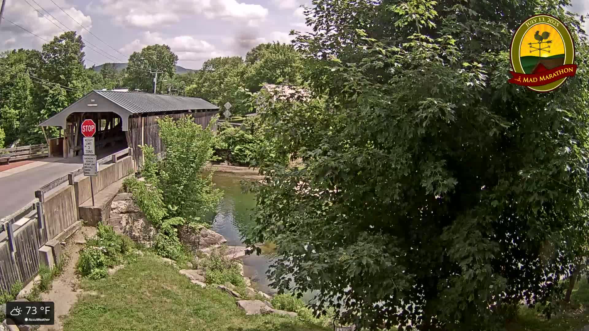 A covered bridge spans a calm river under a partly cloudy sky, surrounded by lush green trees and vegetation.