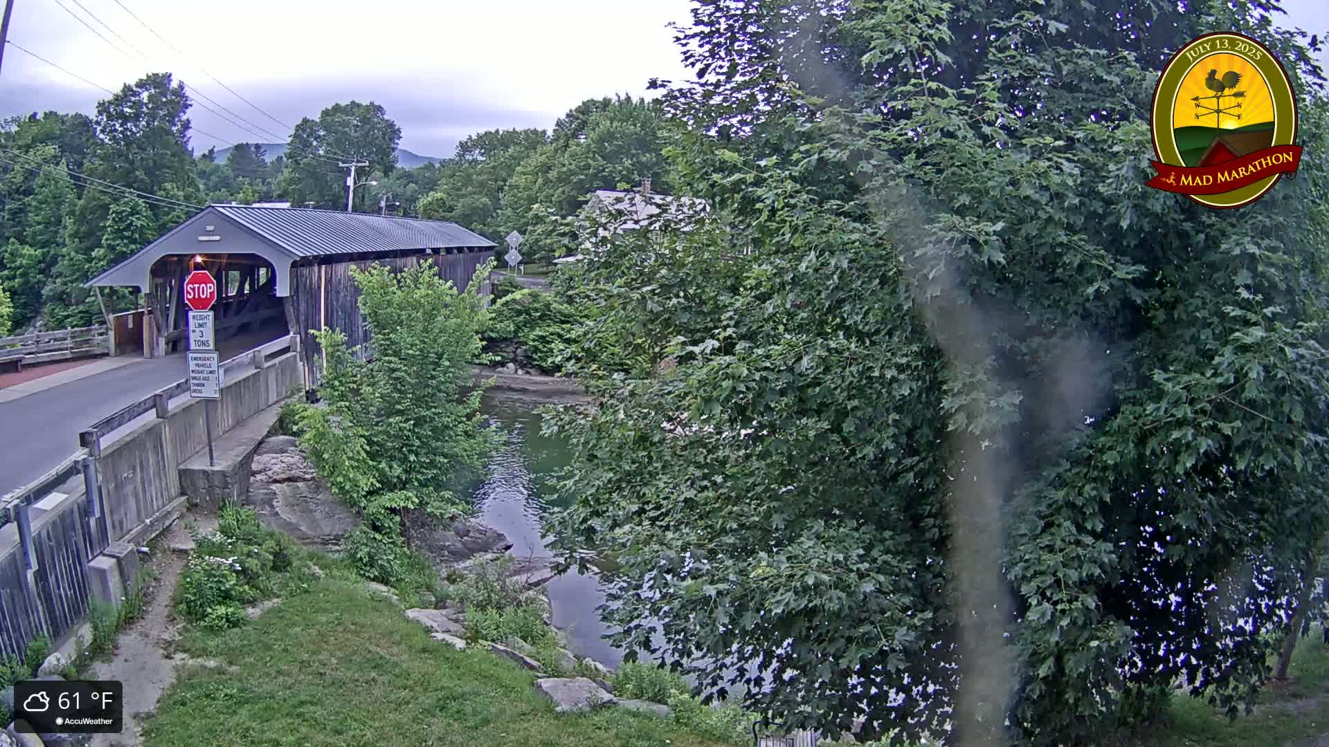 A covered bridge spans a calm river under an overcast sky, surrounded by lush green foliage.