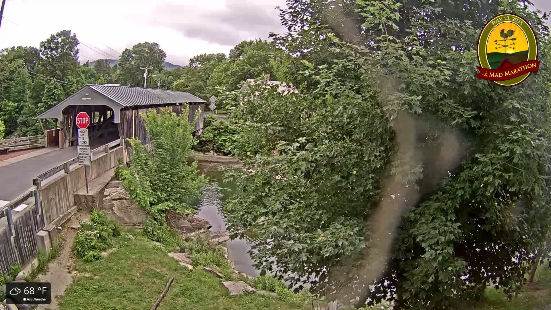 A covered bridge spans a calm river under an overcast sky, surrounded by lush green trees.