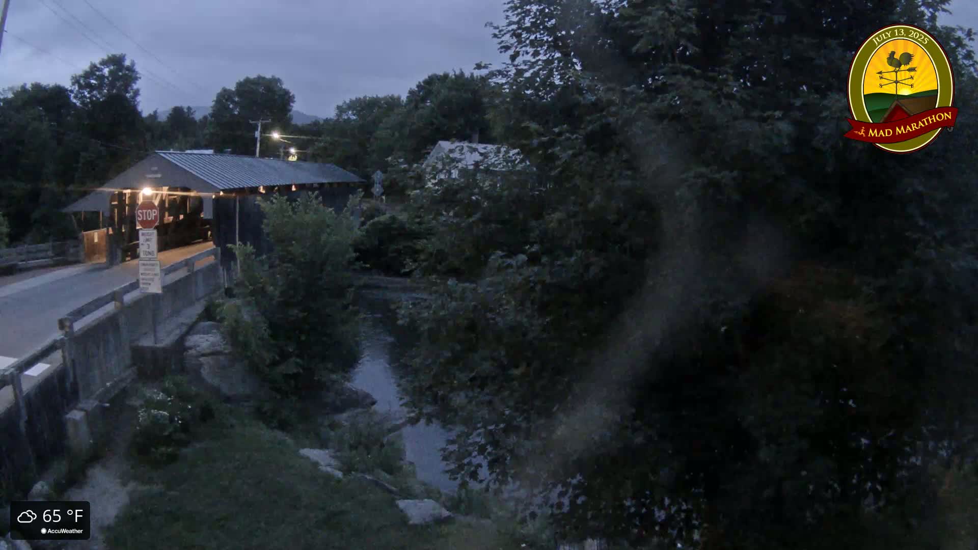 A covered bridge is seen at dusk next to a calm stream, surrounded by lush green trees under a cloudy sky.