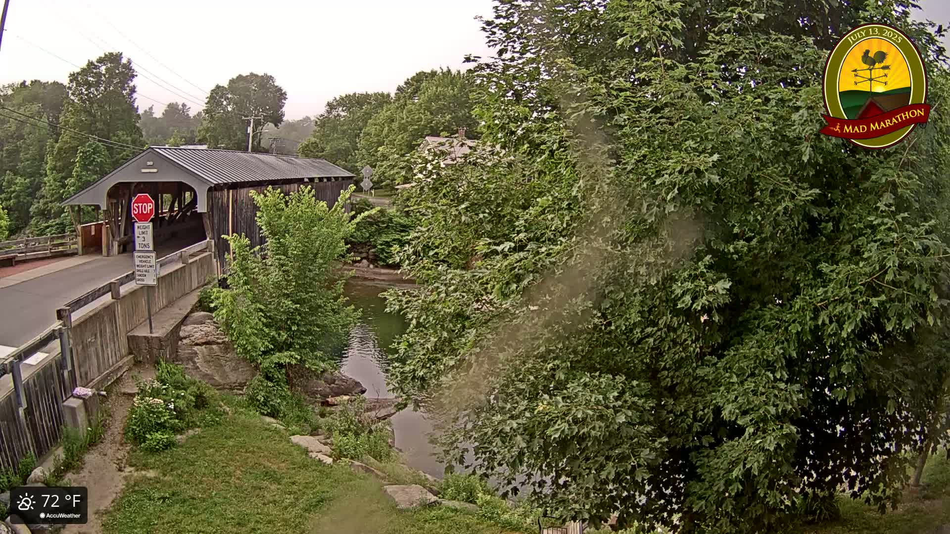 A covered bridge spans a calm river under an overcast sky, surrounded by lush green trees and vegetation.