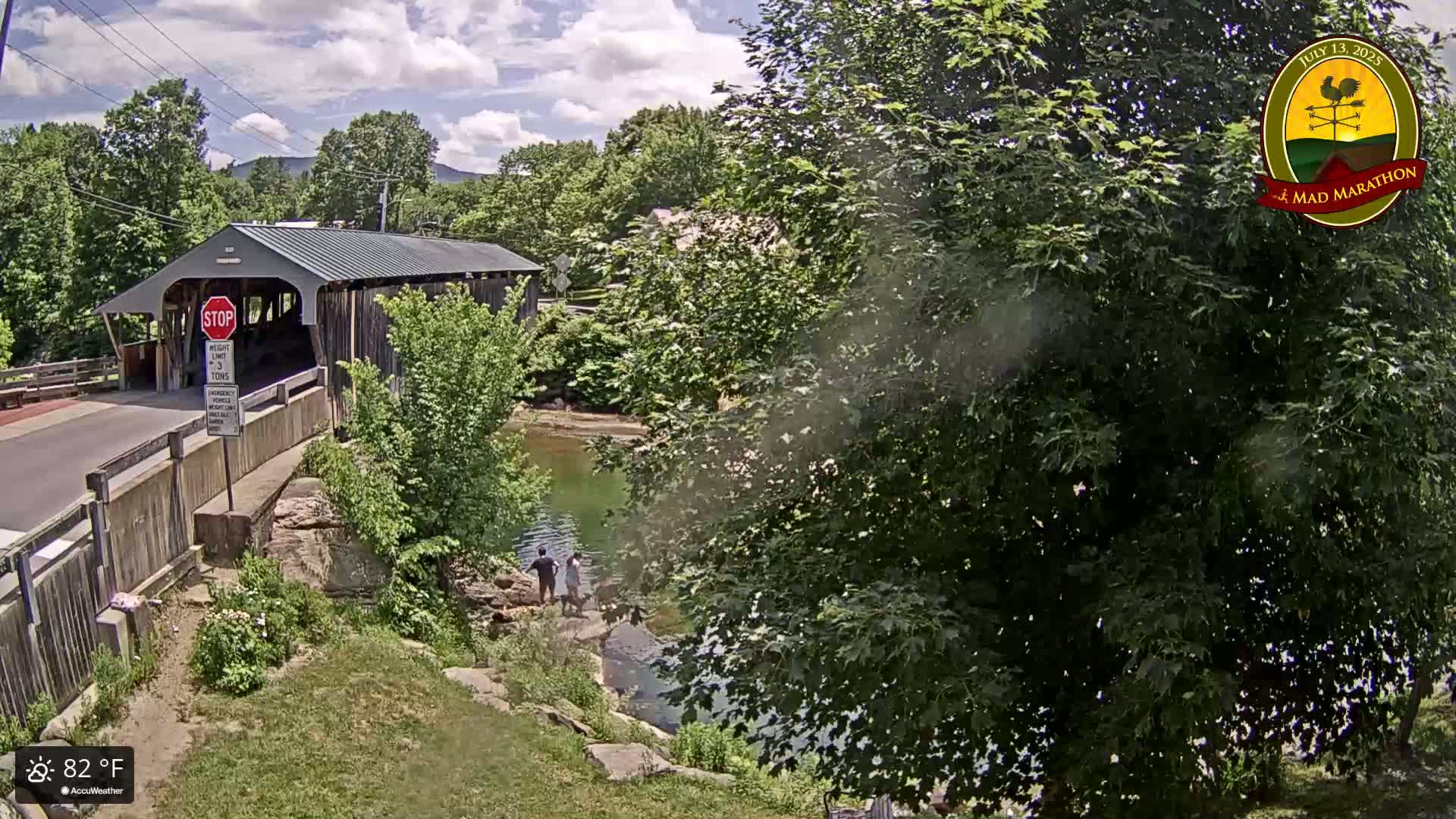 A covered bridge spans a calm river under a partly cloudy sky, with people standing near the water's edge on a sunny 82°F day.