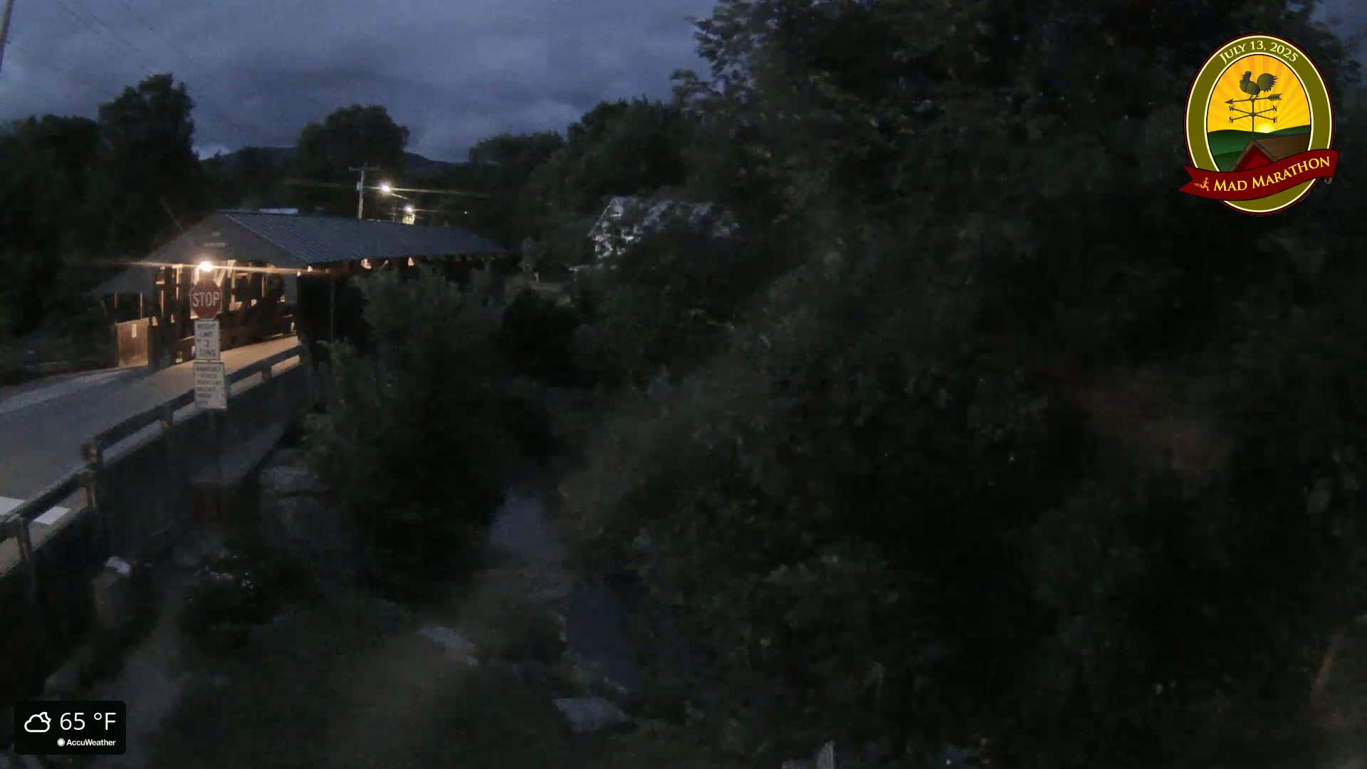 A covered bridge sits over a dark, narrow stream under a twilight sky.