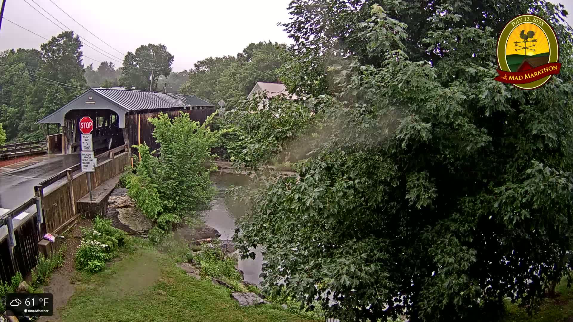 A covered bridge stands over a river under a gray, rainy sky, surrounded by lush green trees and vegetation.