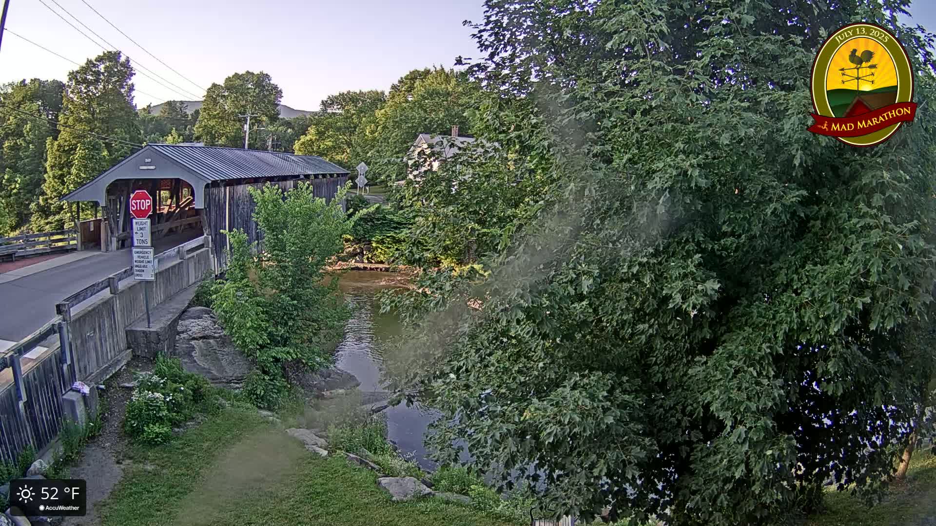 A covered bridge spans a calm river under a clear, mostly sunny sky, surrounded by lush green trees and vegetation.