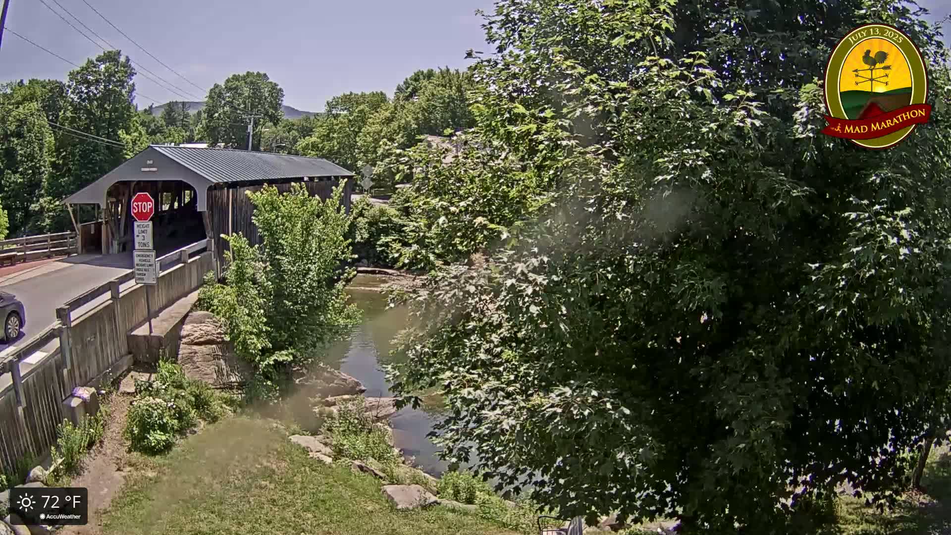 A covered bridge crosses a calm river on a sunny day, surrounded by lush green trees and vegetation.