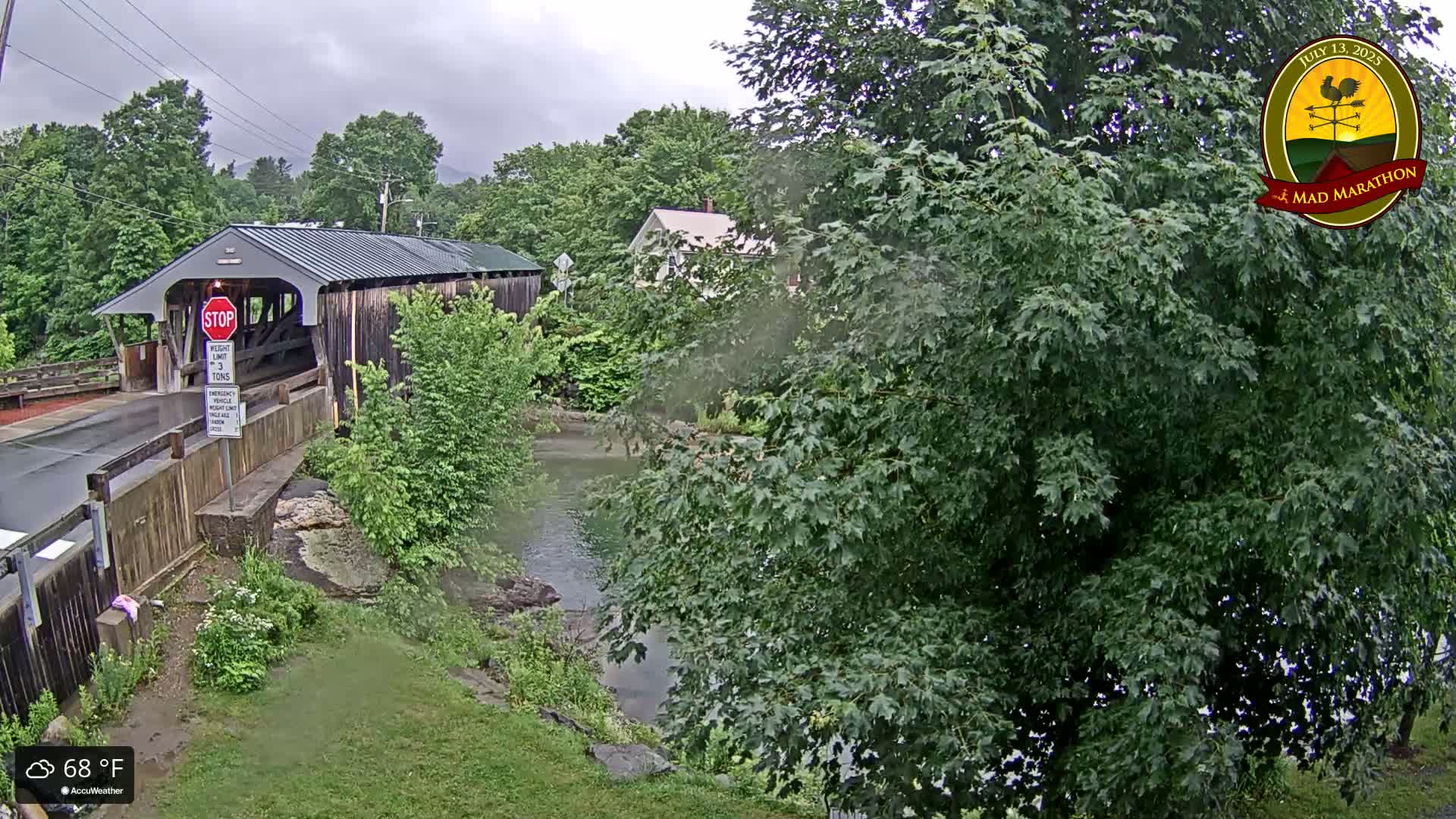 A covered bridge is visible on an overcast day, next to a calm body of water partially obscured by lush green foliage.
