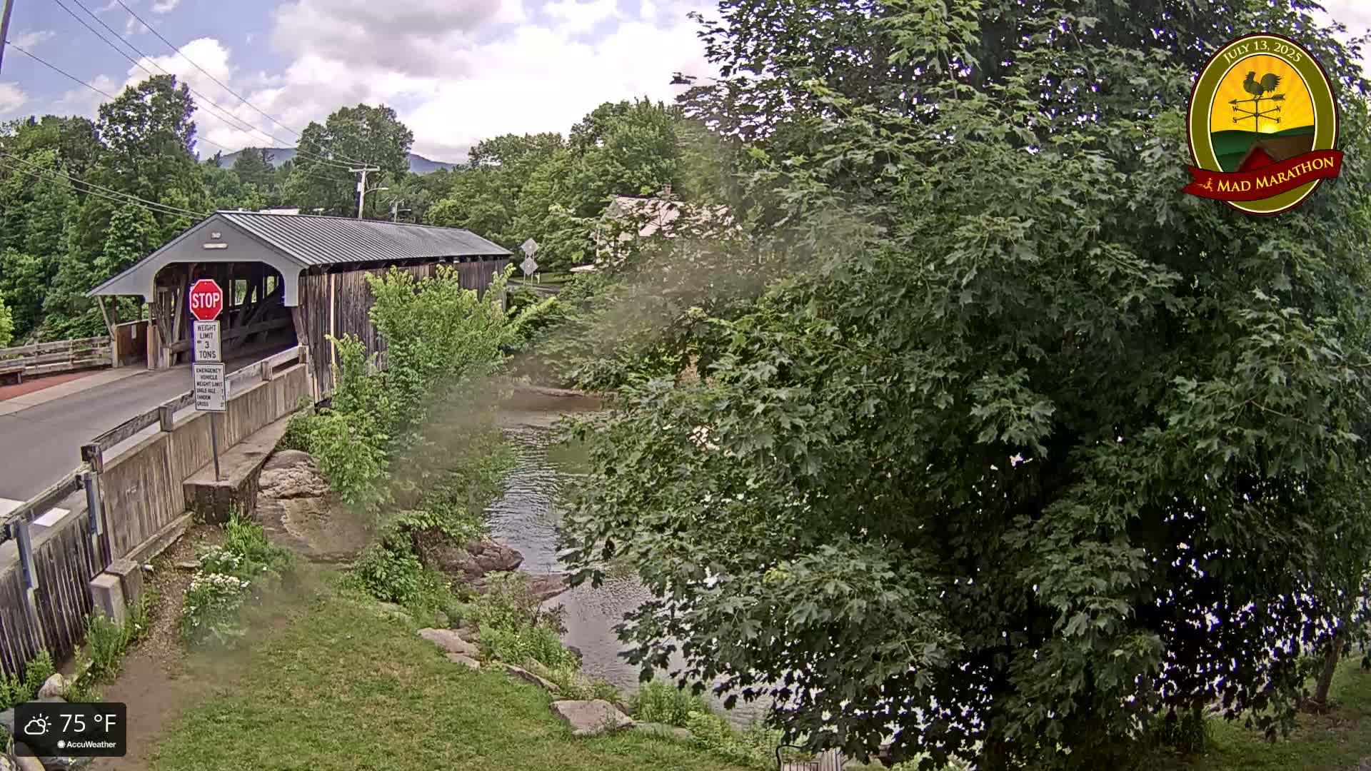 A covered bridge spans a calm river under a partly cloudy sky, surrounded by lush green foliage on a 75°F day.