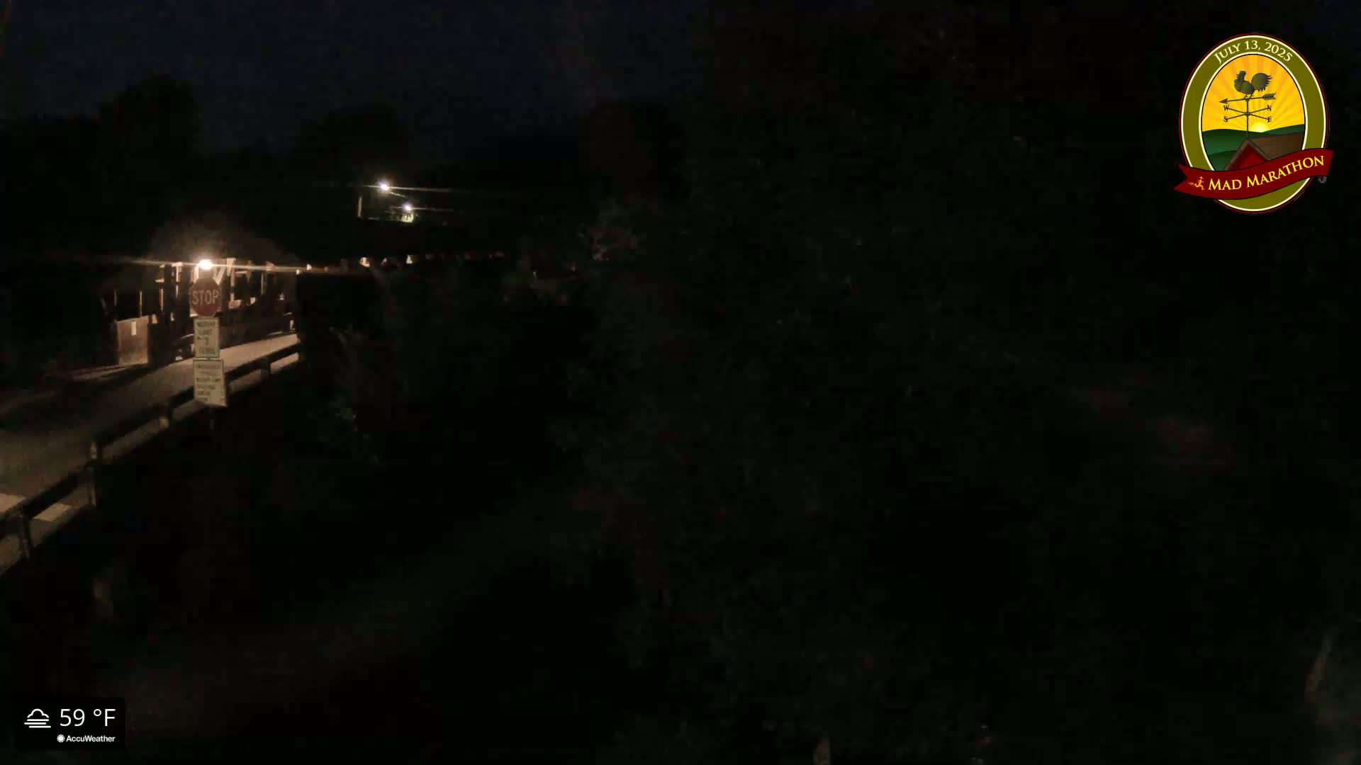 A nighttime, dimly lit view of a bridge with a stop sign, and surrounding dark foliage, under a 59°F sky.