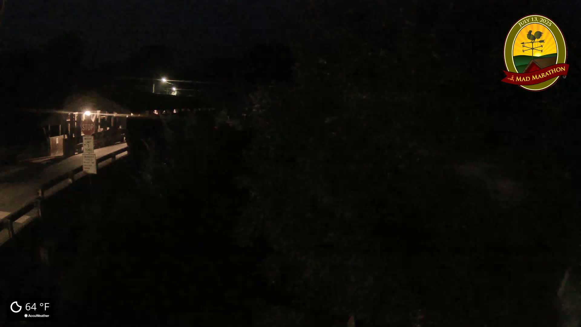 A nighttime, darkly lit view of a covered bridge with a few people visible on the structure, under a clear, 64°F sky.