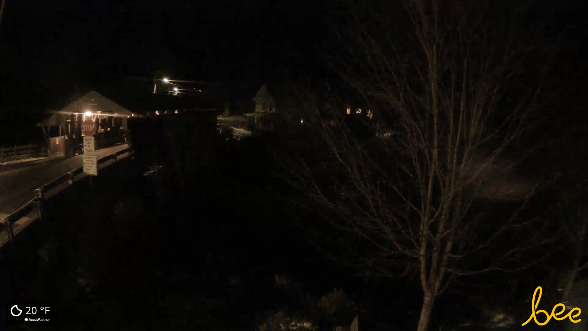 On a cold, dark winter night, an illuminated covered bridge or rustic building is visible on the left, with a bare-branched tree prominent in the right foreground and faint lights from distant structures in the dark landscape.