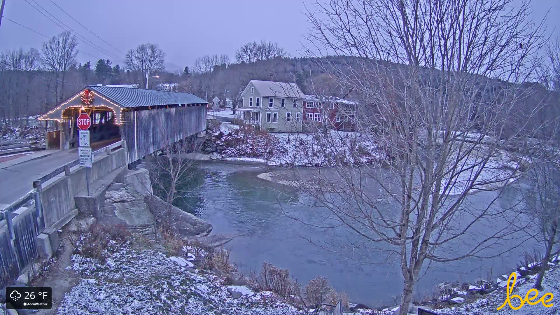 On a cold, dark winter night, an illuminated covered bridge or rustic building is visible on the left, with a bare-branched tree prominent in the right foreground and faint lights from distant structures in the dark landscape.