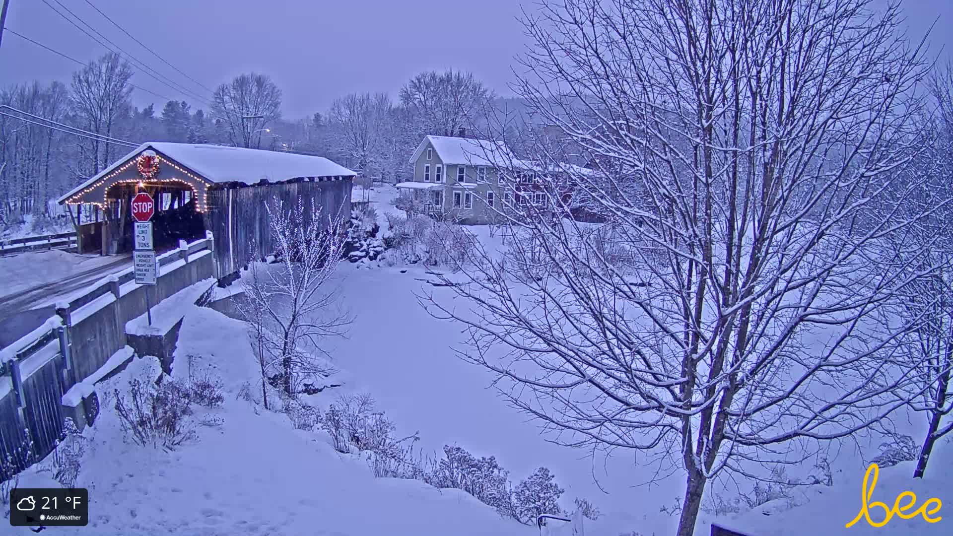 On a cold, dark winter night, an illuminated covered bridge or rustic building is visible on the left, with a bare-branched tree prominent in the right foreground and faint lights from distant structures in the dark landscape.