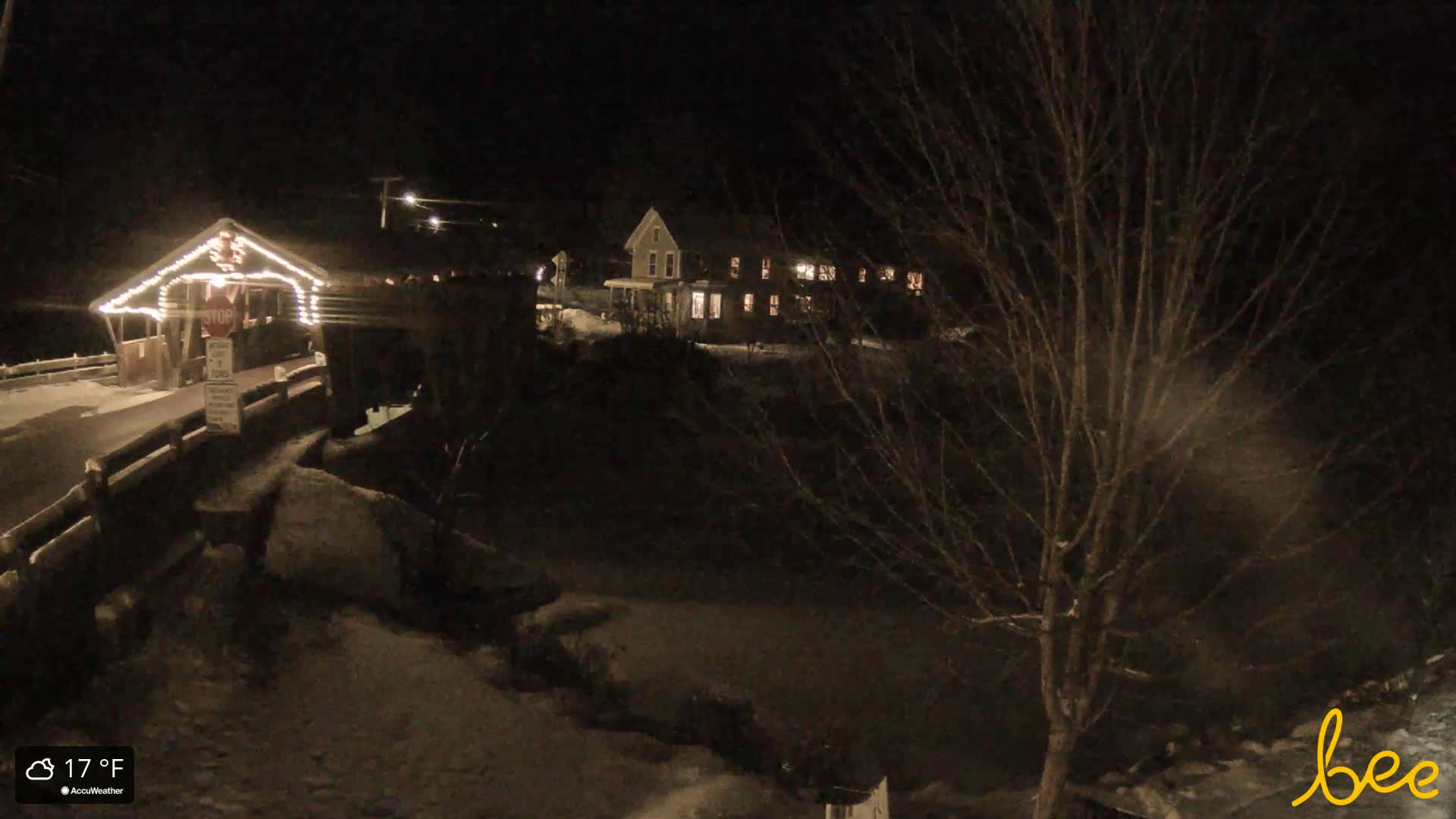 On a cold, dark winter night, an illuminated covered bridge or rustic building is visible on the left, with a bare-branched tree prominent in the right foreground and faint lights from distant structures in the dark landscape.