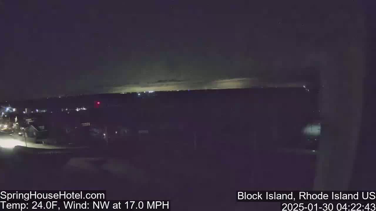 A nighttime, wide shot of a town and distant horizon under a mostly clear, dark sky with a faint glow on the horizon, and a northwest wind at 17 mph and a temperature of 24°F.