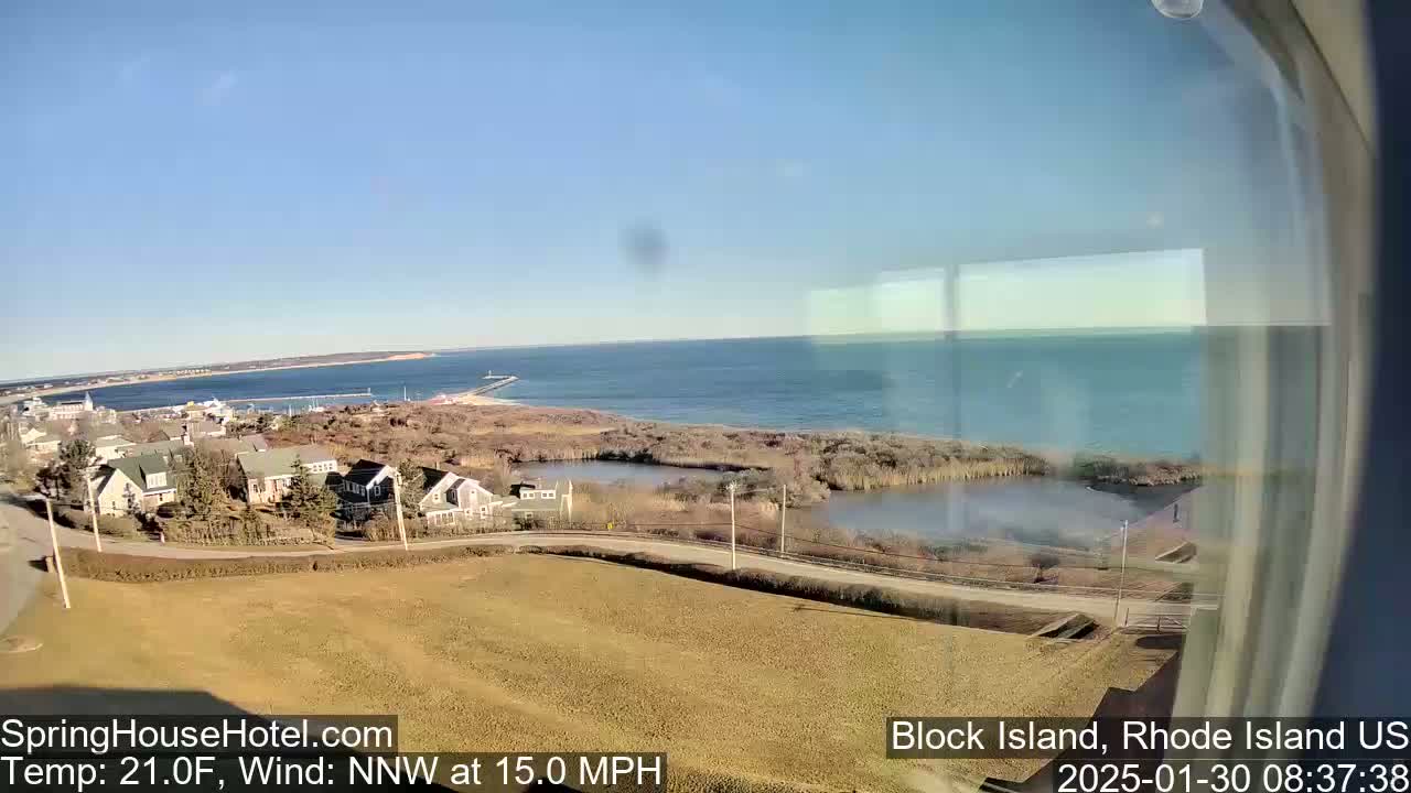 A coastal view shows a calm ocean under a clear blue sky, with houses and marshland in the foreground, and a 15 mph northwest wind at 21 degrees Fahrenheit.