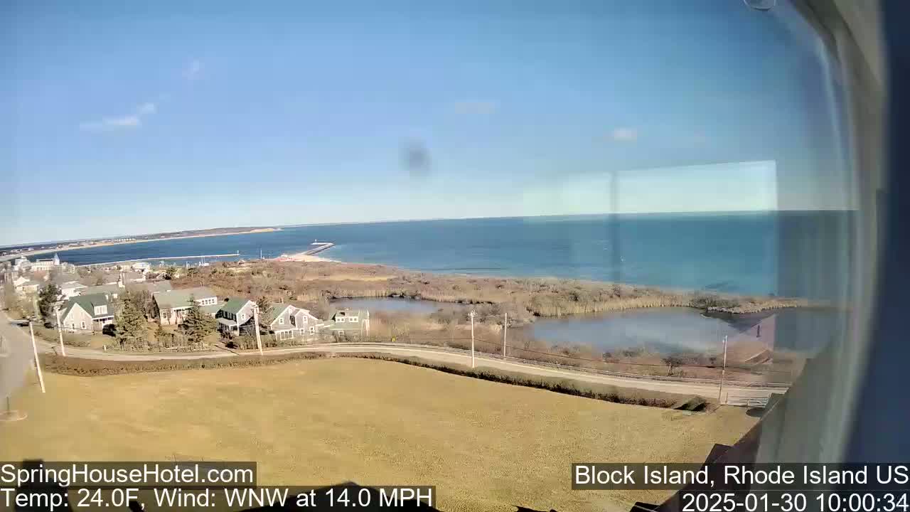 A coastal view on a sunny day shows houses, a road, marshland, and a body of water extending to the horizon.