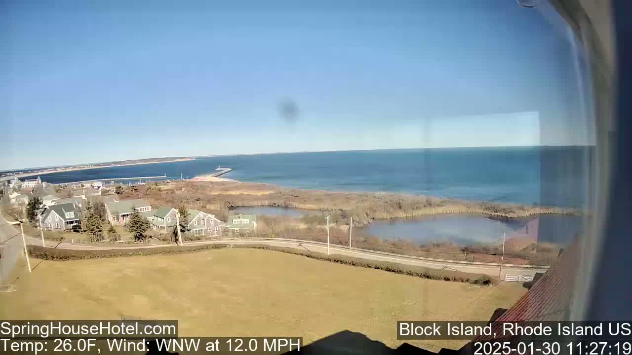 A coastal scene on a clear, sunny day shows a residential area overlooking a calm body of water and a distant shoreline, with a 12 mph wind.
