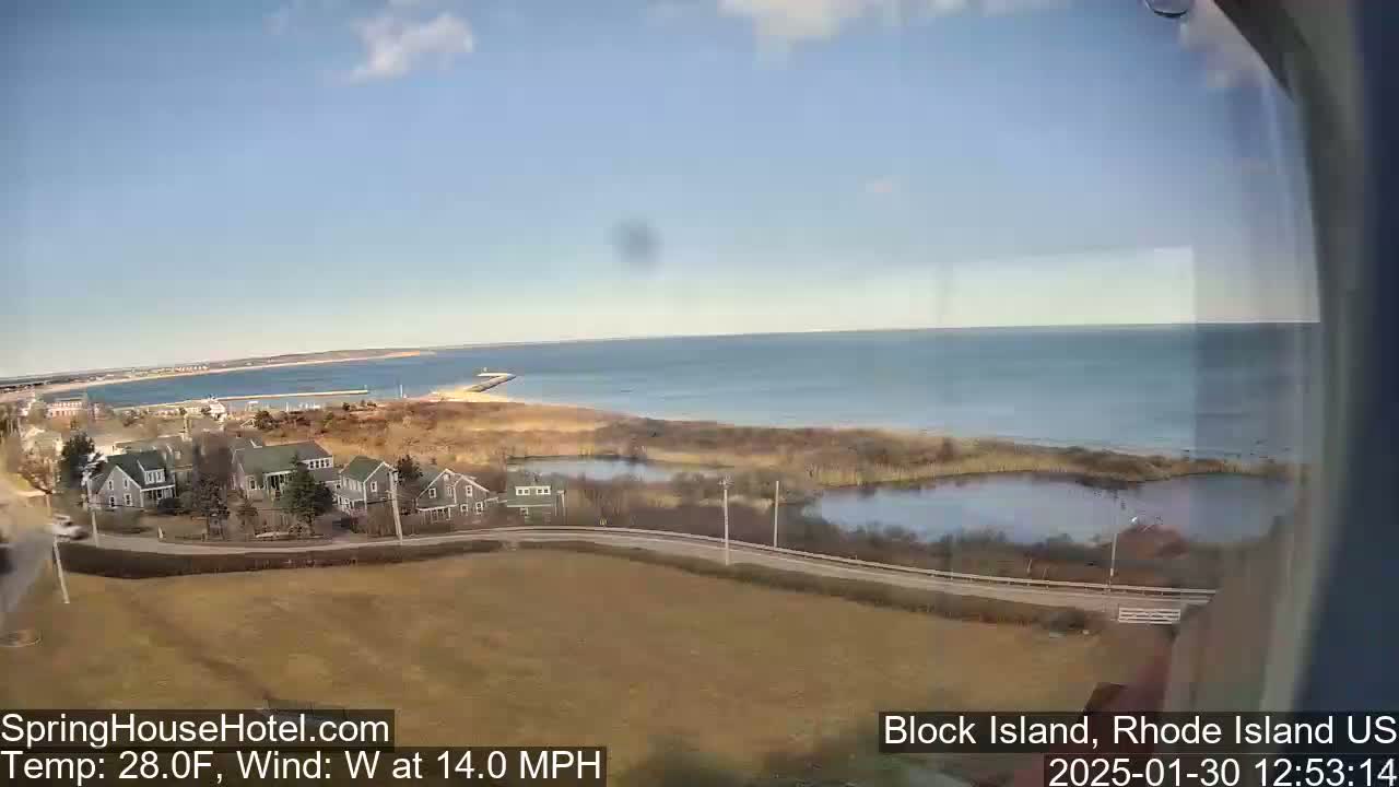 A coastal view on a sunny day shows a calm body of water, a sandy shore, and a cluster of houses, with a 28°F temperature and a 14 mph westerly wind.