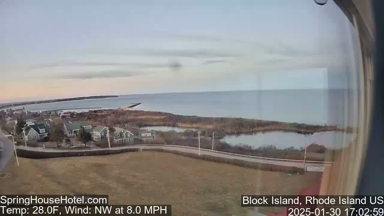 A coastal view shows a calm ocean under a pale sky, with houses and marshland in the foreground, and a wind of 8 mph from the northwest.
