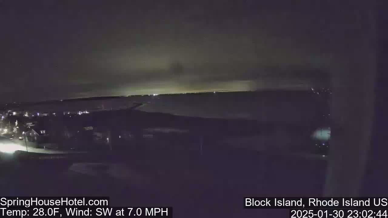 A nighttime, elevated view of a dark, coastal town with a calm body of water in the background, under a cloudy sky, with a southwest wind at 7 mph and a temperature of 28°F.