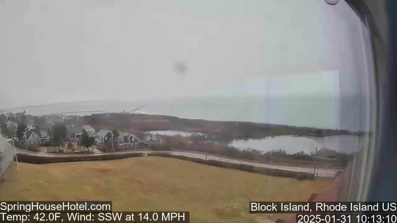 A coastal scene with houses and a calm body of water is visible under an overcast sky, with a wind blowing from the south-southwest at 14 miles per hour.
