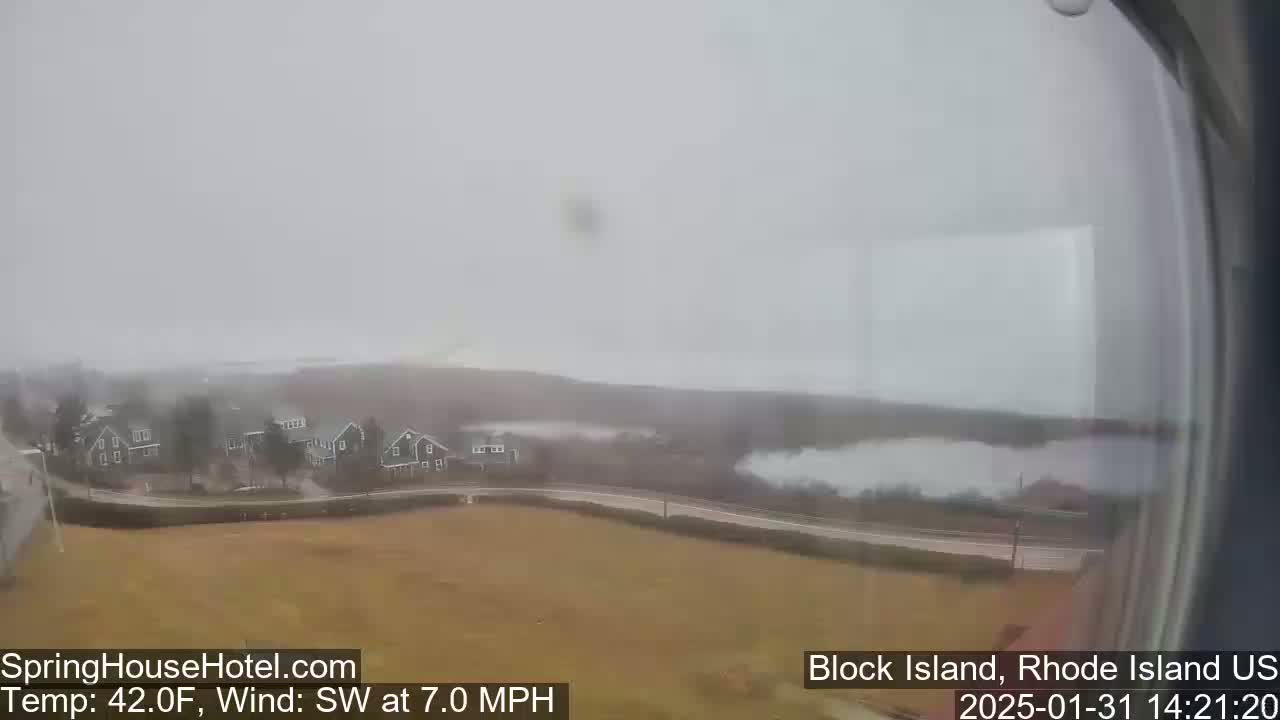 A hazy, overcast view of a coastal town with houses, a road, and a body of water in the distance, under a 42°F temperature with a 7.0 mph southwest wind.