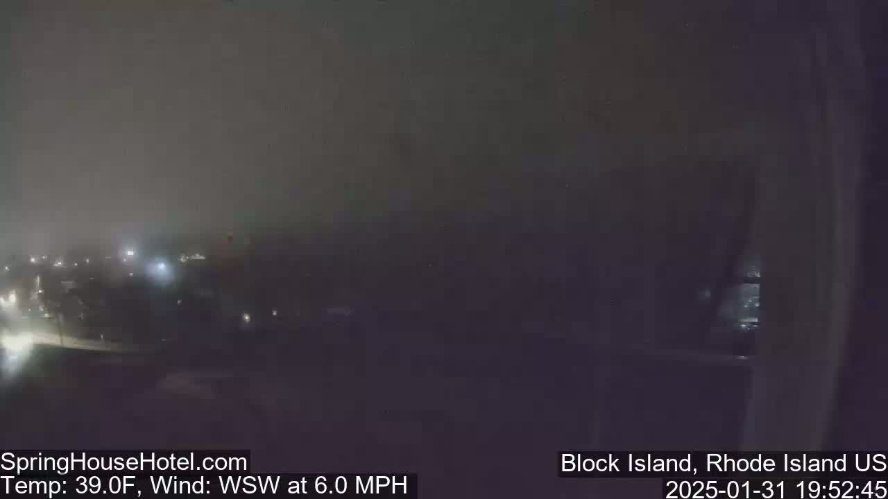 A hazy, nighttime view of a town with faint lights, under a dark sky and a WSW wind of 6.0 MPH at 39°F.