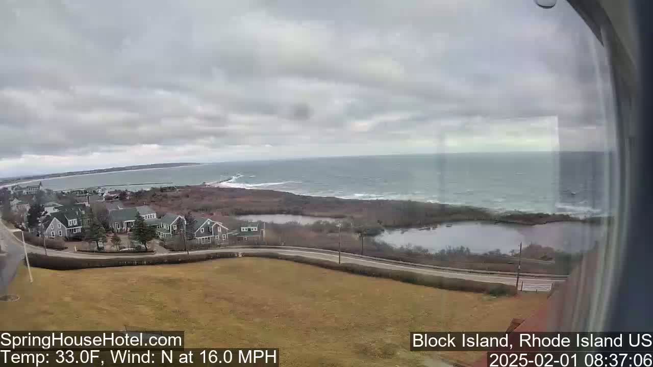 A coastal view on a cloudy day shows a calm ocean, a road winding through houses near a shoreline, and a small pond inland.