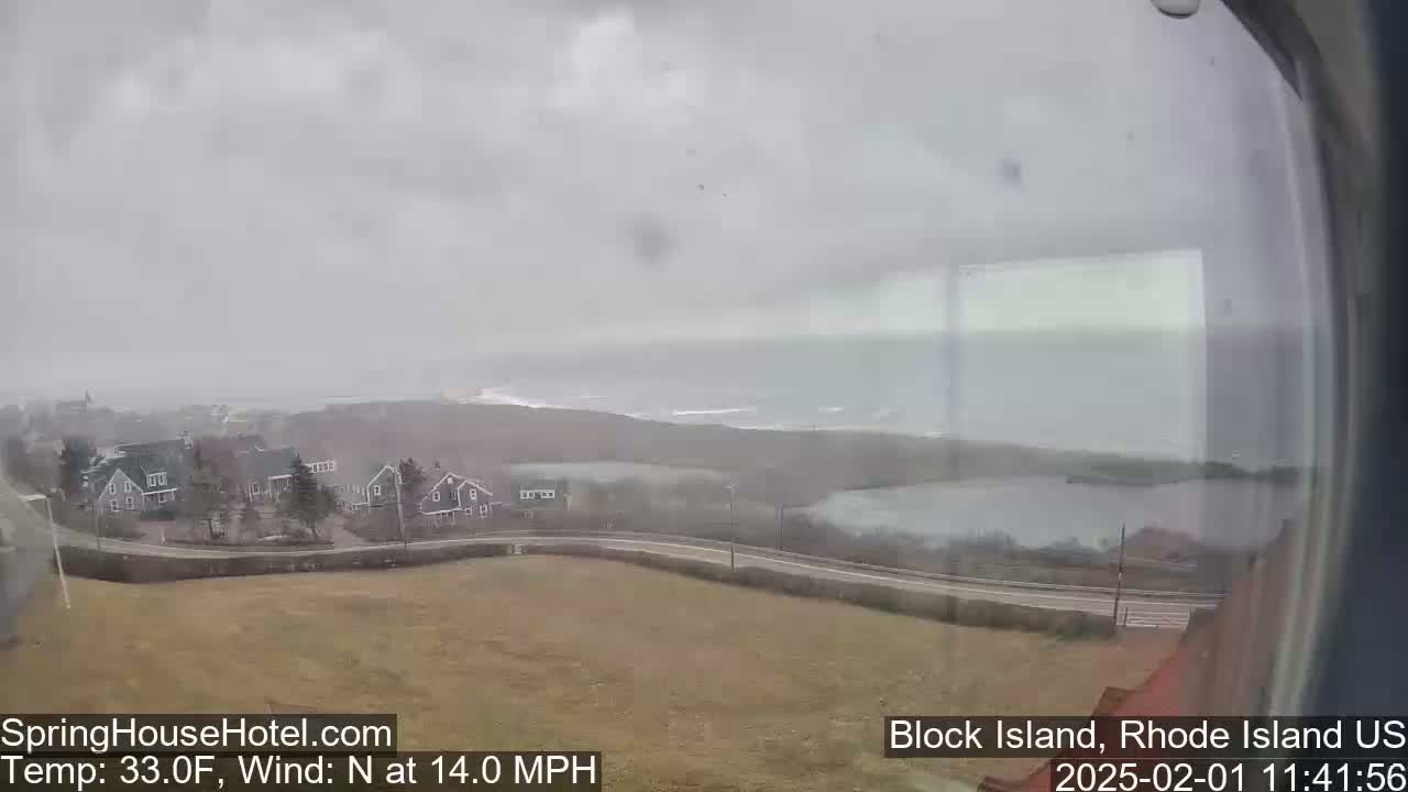 A coastal view shows a gray, overcast sky, houses, a road, a body of water, and a stretch of coastline under windy conditions.