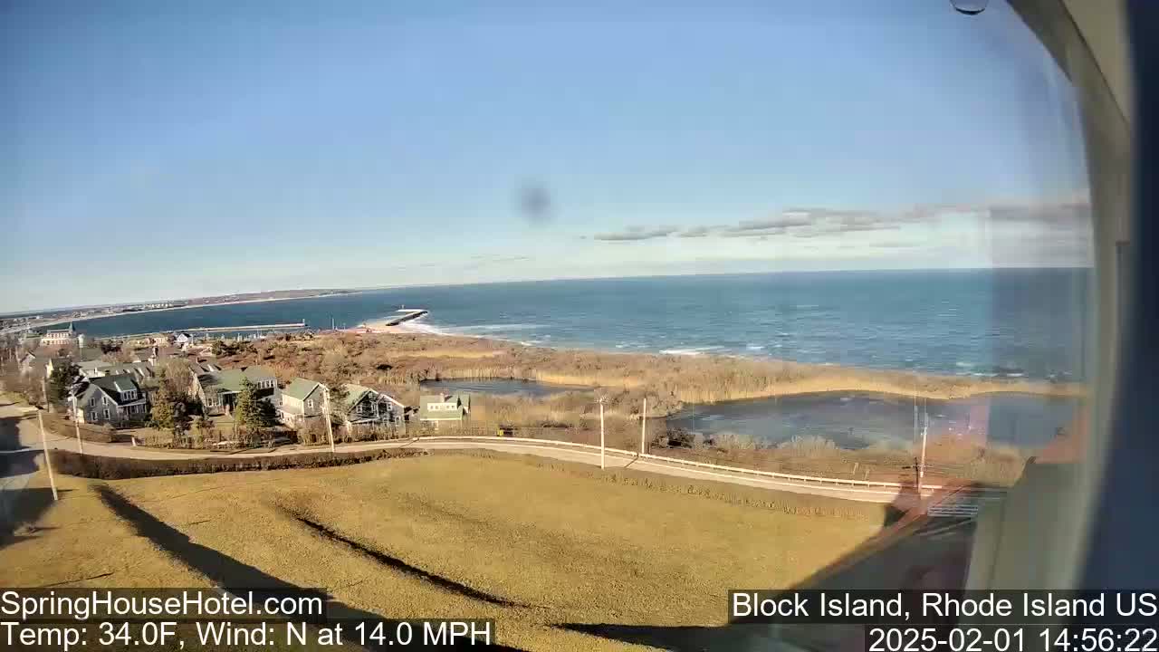 A coastal view on a partly sunny day shows a calm ocean with houses and marshland in the foreground, under a  north wind of 14 mph and a temperature of 34°F.
