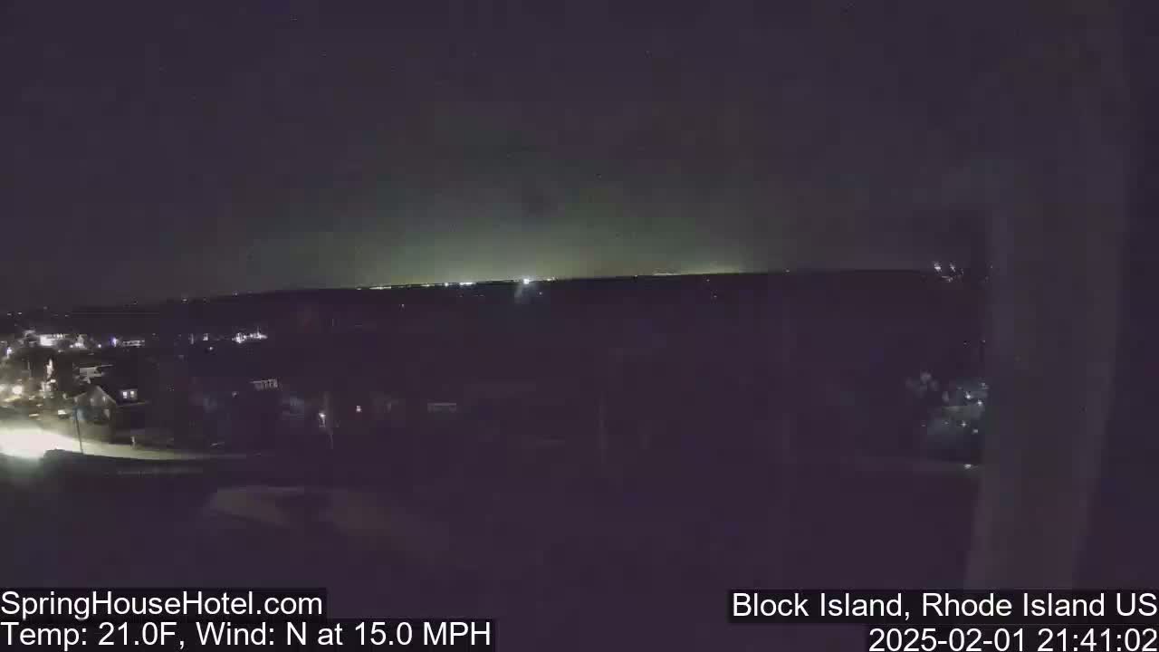 A nighttime view of a town illuminated by streetlights, with a dark horizon under a clear sky and a north wind blowing at 15 mph.
