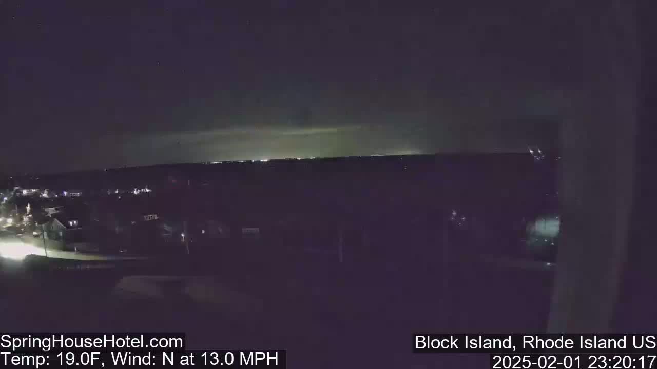 A nighttime view of a town with a distant, bright horizon under a dark, clear sky with a north wind at 13 mph.