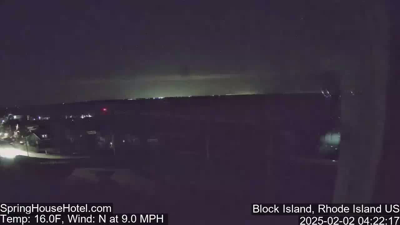 A nighttime, elevated view of a town illuminated by lights, with a calm, dark sky and a slight north wind.