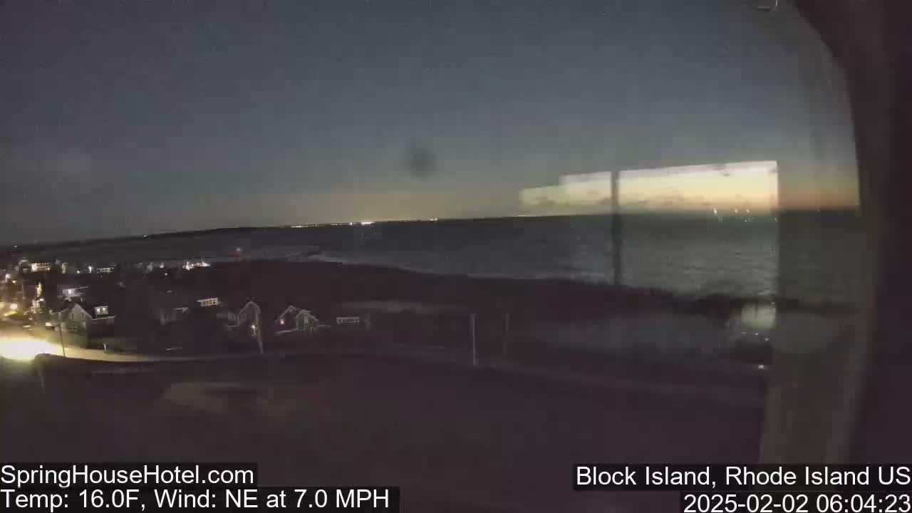 A nighttime view of a dark ocean under a mostly clear sky, with a small town visible on the left and a slight northeastern wind.