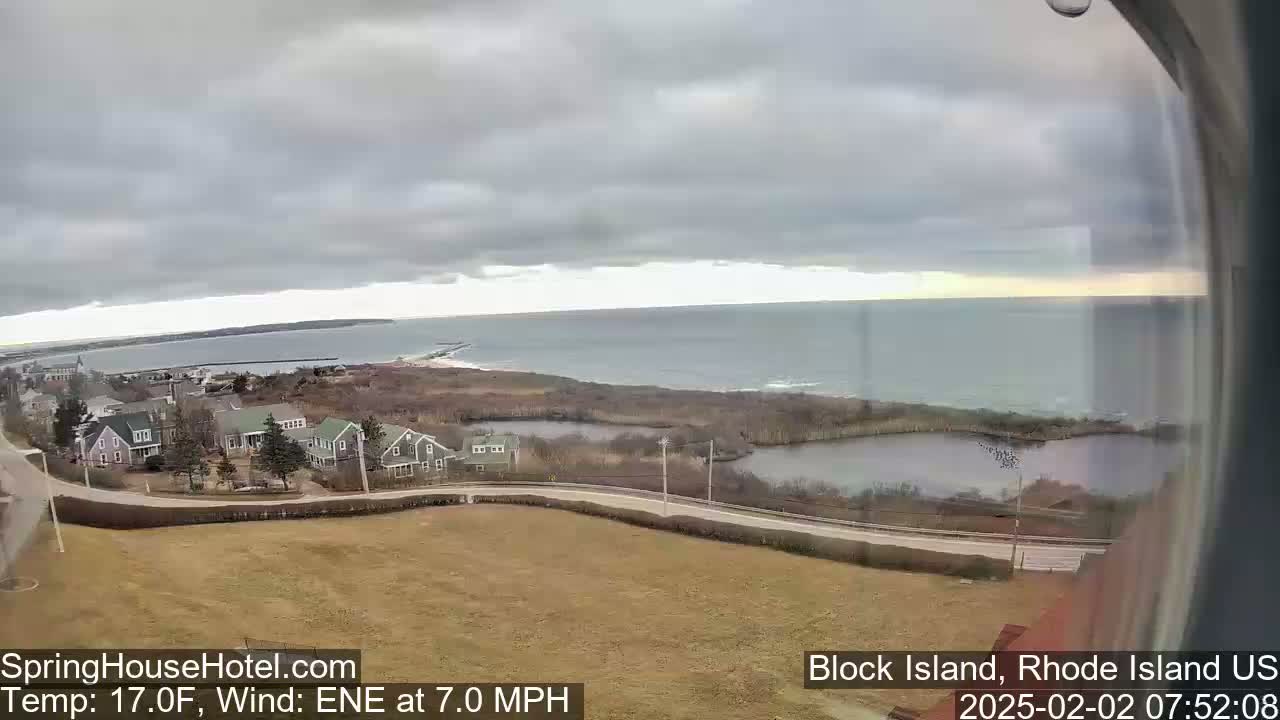 A coastal view shows a calm ocean under an overcast sky, with houses and a road visible in the foreground.