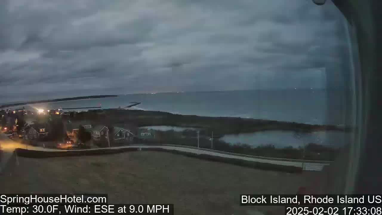 A coastal scene at dusk shows a calm ocean, a dark sky, and a few houses with lights on near a road.