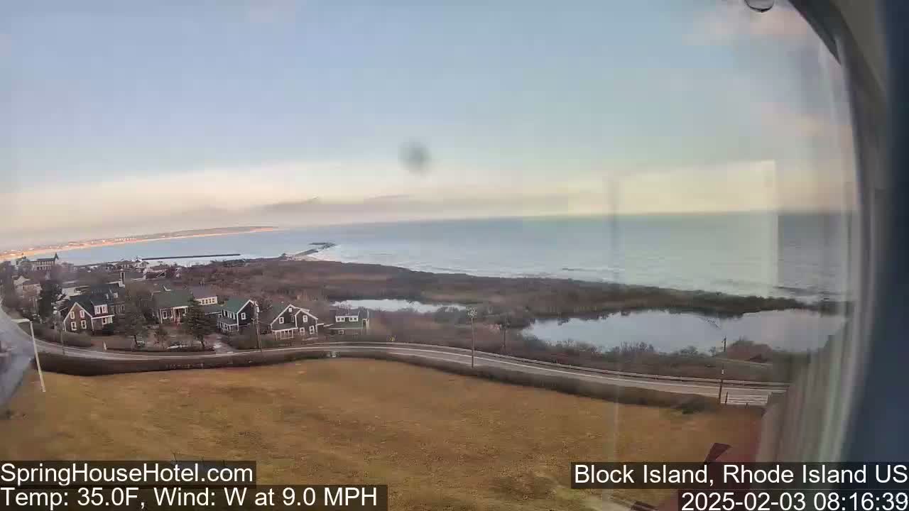 A coastal view shows a calm ocean under a mostly clear sky, overlooking houses and a marshy area with a road in the foreground.