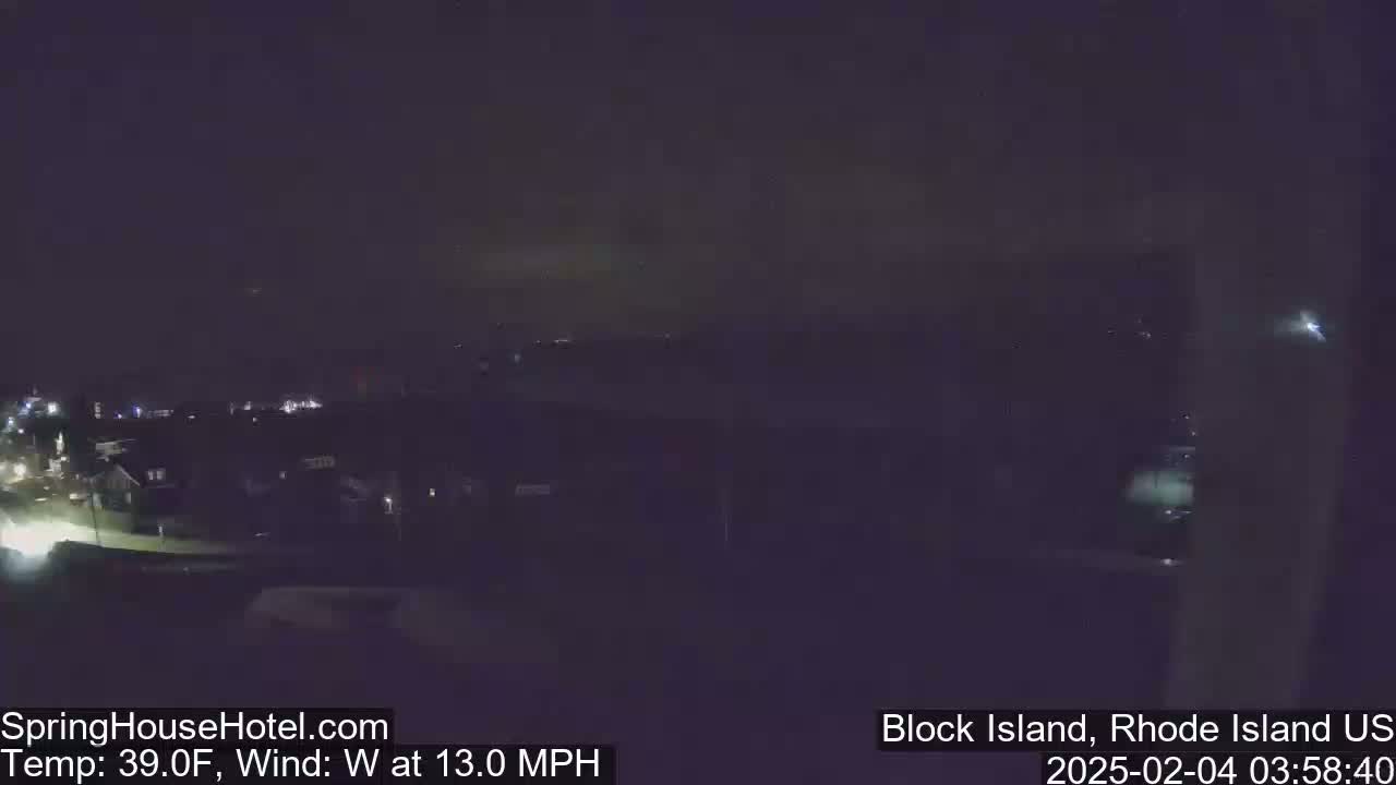 A nighttime long shot of a town shows dimly lit houses and streets under a dark sky with a westerly wind at 13 mph and a temperature of 39°F.