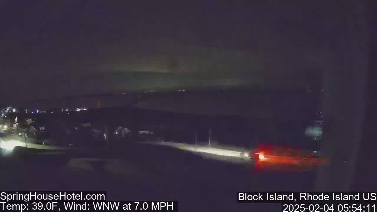 A nighttime, low-light view of a road with a few houses visible in the distance under a dark sky with a faint greenish glow near the horizon, a 7 mph wind from the west-northwest, and a temperature of 39°F.